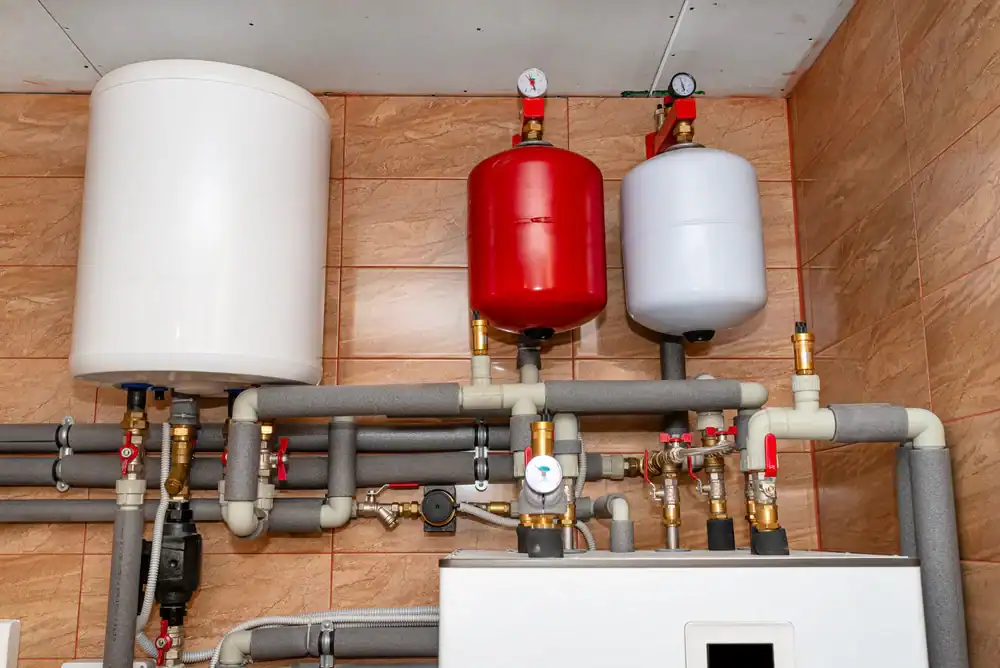 Water heating system with connected pipes and valves, featuring a large white tank on the left, a red expansion tank in the center, and a smaller white tank on the right—professionally installed by a plumber Brevard County, FL, against a tiled wall.