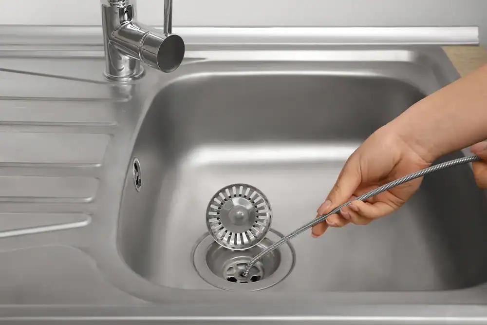 A hand uses a flexible drain snake to clean a stainless steel kitchen sink in Brevard County, FL, with the sink strainer lifted out&mdash;just like a professional plumber would do.