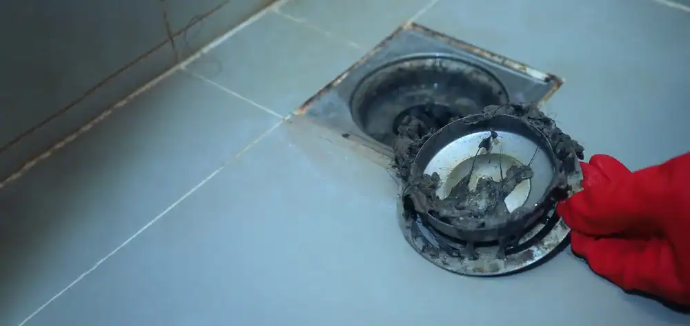 A hand in a red glove holds a dirty, clogged drain cover over a tiled floor next to an open drain, showing accumulated debris&mdash;a reminder that calling a plumber Brevard County FL can help prevent stubborn blockages like these.