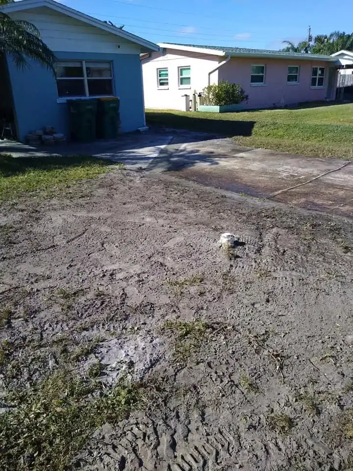 A patch of bare dirt with sparse grass covers the front yard of a blue house; a short white pipe, possibly left by a plumber in Brevard County, FL, sticks up from the ground near a concrete driveway. Another house is visible in the background.