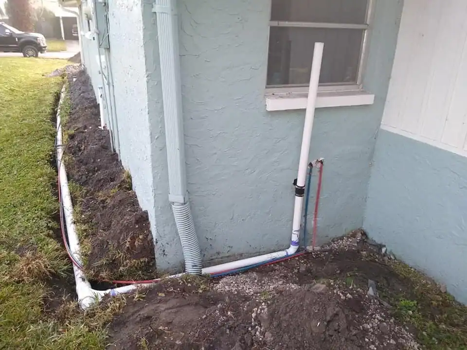 White PVC and flexible pipes run along the base of a light blue FL house, partially buried in soil with fresh dirt visible, near a window and the corner—work typical of a skilled plumber Brevard County residents trust.