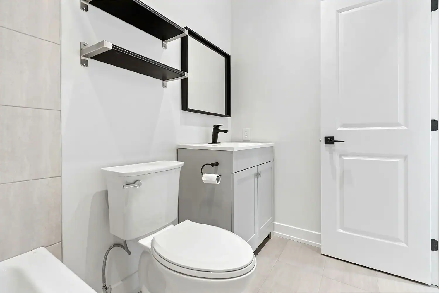 Modern bathroom with white walls, gray vanity and black faucet, rectangular mirror, two black shelves, and a white door. Light tile flooring and partial view of a bathtub—expertly maintained by a plumber Brevard County, FL trusts.