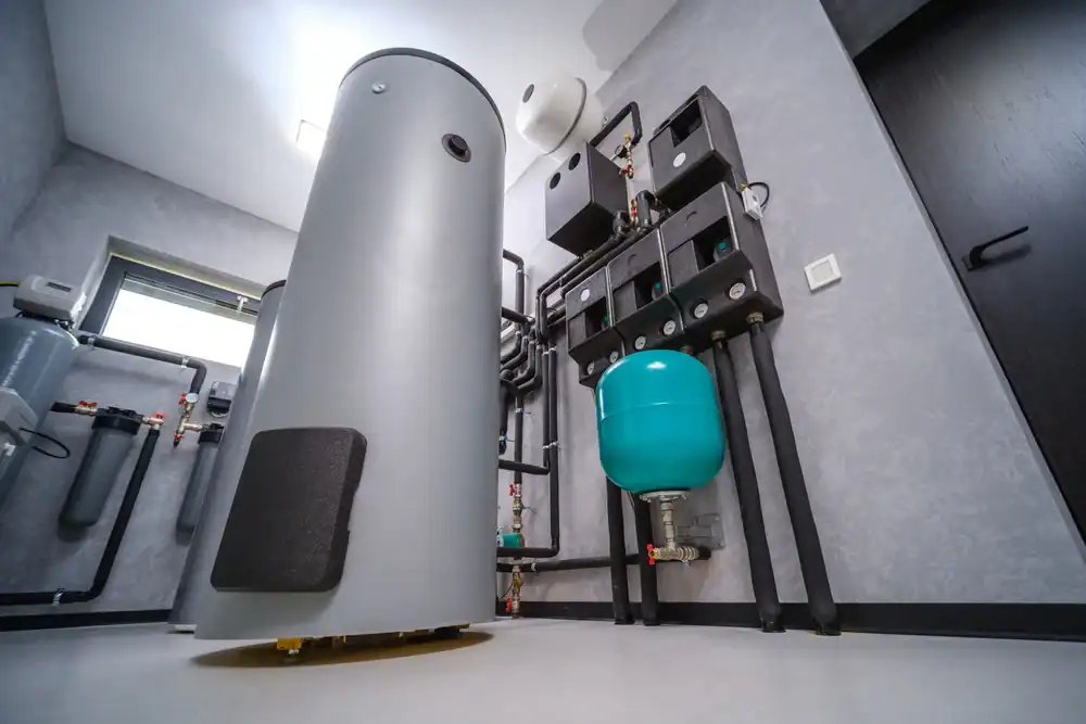 Modern utility room with a large cylindrical water heater, blue expansion tank, wall-mounted control panels, and pipes on gray walls and floor. A window and black door complete the space&mdash;ideal work for a skilled plumber in Brevard County, FL.