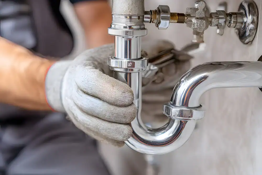 A plumber Brevard County, FL, wearing gray gloves, is installing or repairing a shiny chrome pipe under a sink, with tools and valves visible on the wall in the background.