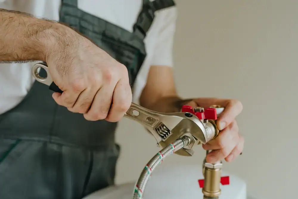 A plumber in Brevard County, FL, wearing work overalls, uses a wrench to tighten plumbing fittings on metal pipes with red handles. The background is plain and out of focus.