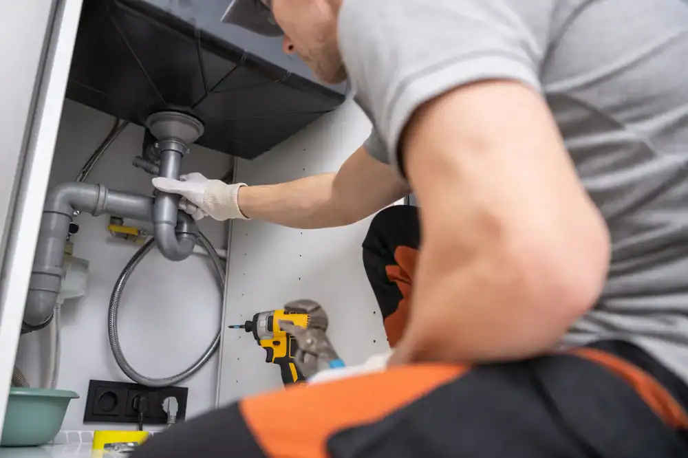 A plumber wearing gloves kneels under a sink, inspecting gray pipes. A yellow drill and pliers are beside him, with a green bowl and electrical outlet in the background.