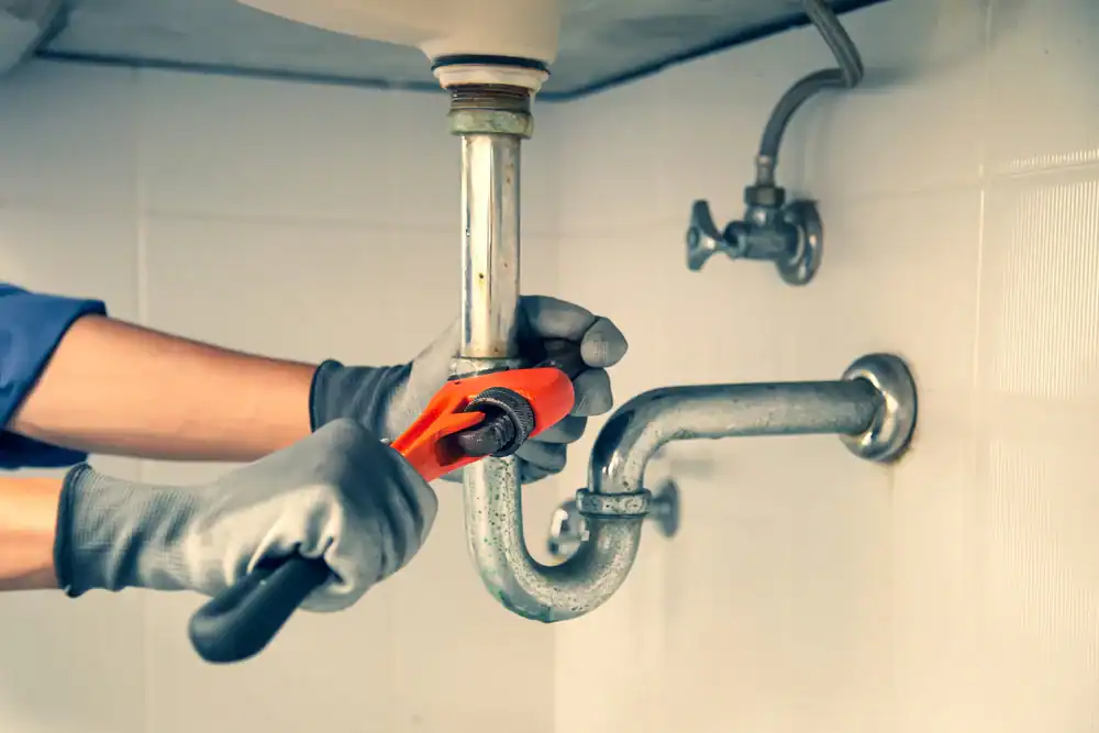 A person wearing gray gloves uses a red pipe wrench to tighten or loosen the piping under a sink in a bathroom or kitchen.