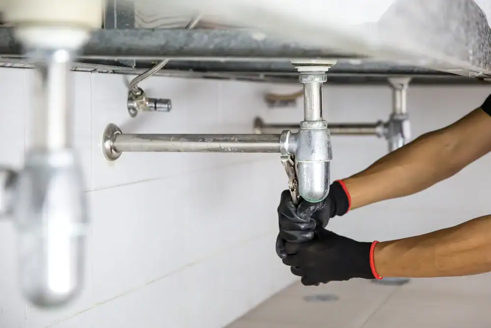 A plumber Brevard County in FL, wearing black gloves, uses a wrench to tighten or loosen a pipe under a sink, performing plumbing maintenance or repair work.