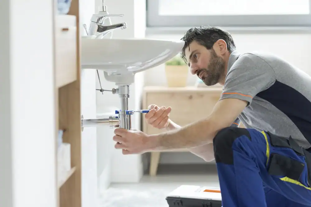 A plumber Brevard County professional in workwear uses a wrench to fix pipes under a bathroom sink. A toolbox sits on the floor, while a plant rests on a wooden table in the background of this FL home.