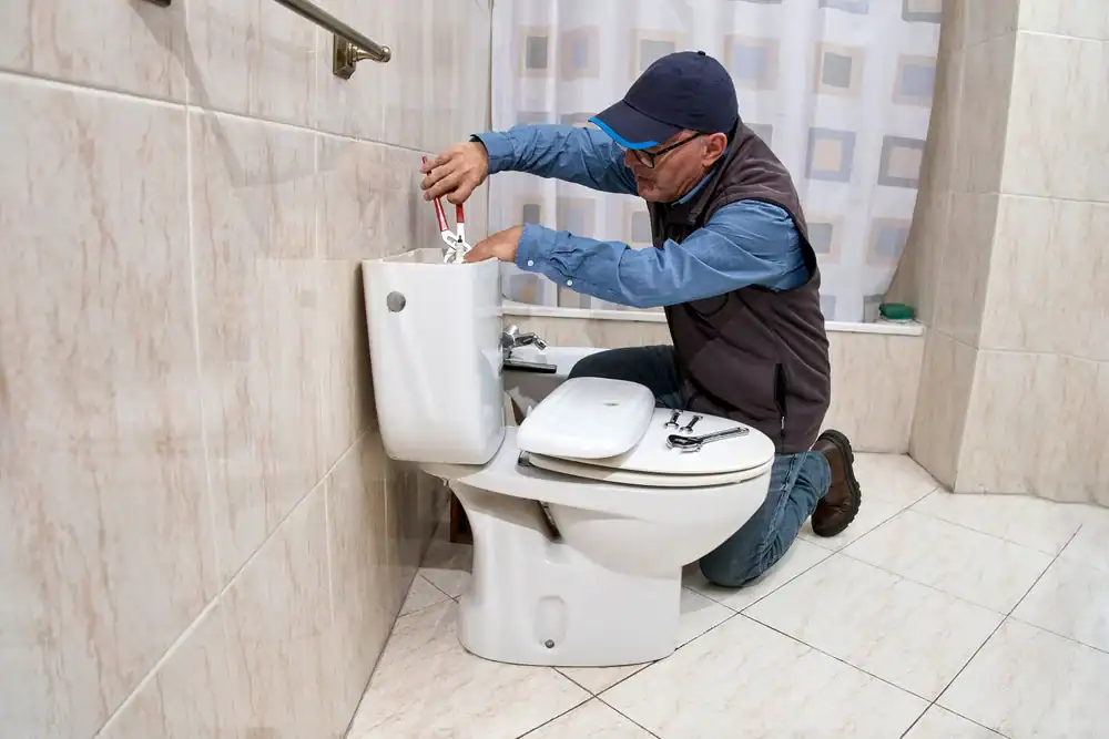 A plumber from Brevard County, FL, wearing a cap and glasses, kneels next to a toilet, using a wrench to repair the tank inside a tiled bathroom.