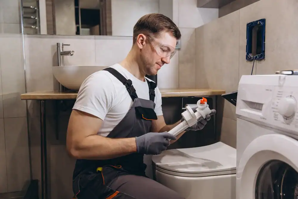 A man wearing work overalls, gloves, and safety glasses is fixing a plumbing part in a bathroom, sitting next to a washing machine and toilet, with a sink in the background.