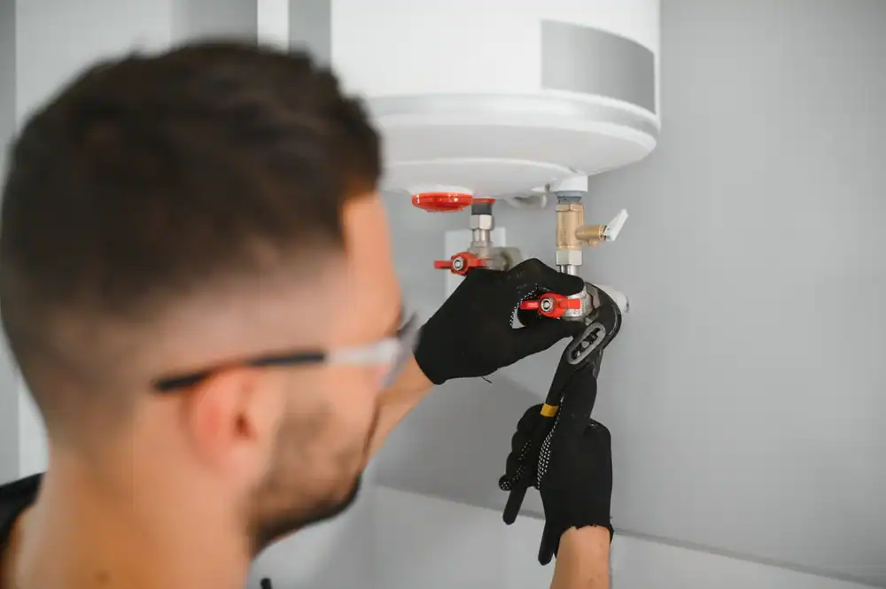 A plumber Brevard County wearing black gloves uses a wrench to adjust pipes connected to a white water heater mounted on a wall.