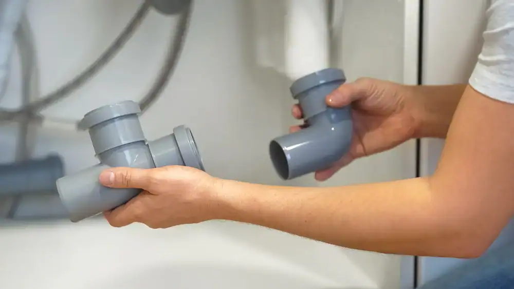 A person holding and assembling two gray plastic plumbing pipes under a sink, preparing to connect them.