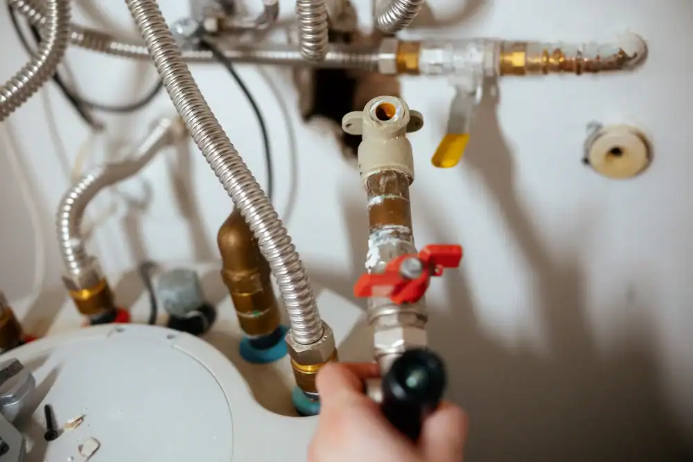 A close-up of a person’s hand adjusting a valve on a network of metal pipes connected to a water heater, with various connectors, valves, and flexible hoses visible.