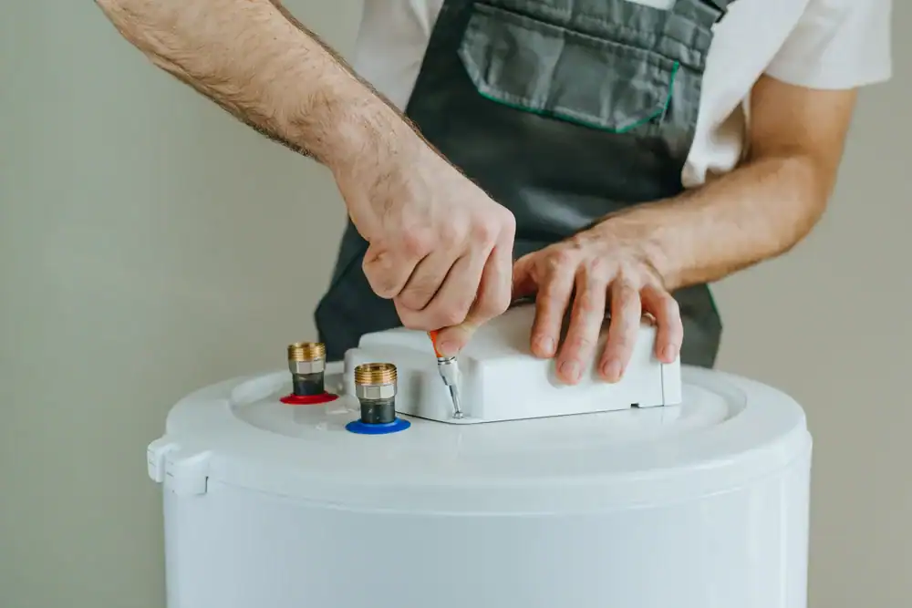 A plumber in Brevard County, FL, wearing work overalls, uses a screwdriver to repair or install a white water heater, focusing on the connectors and wiring at the top of the appliance.