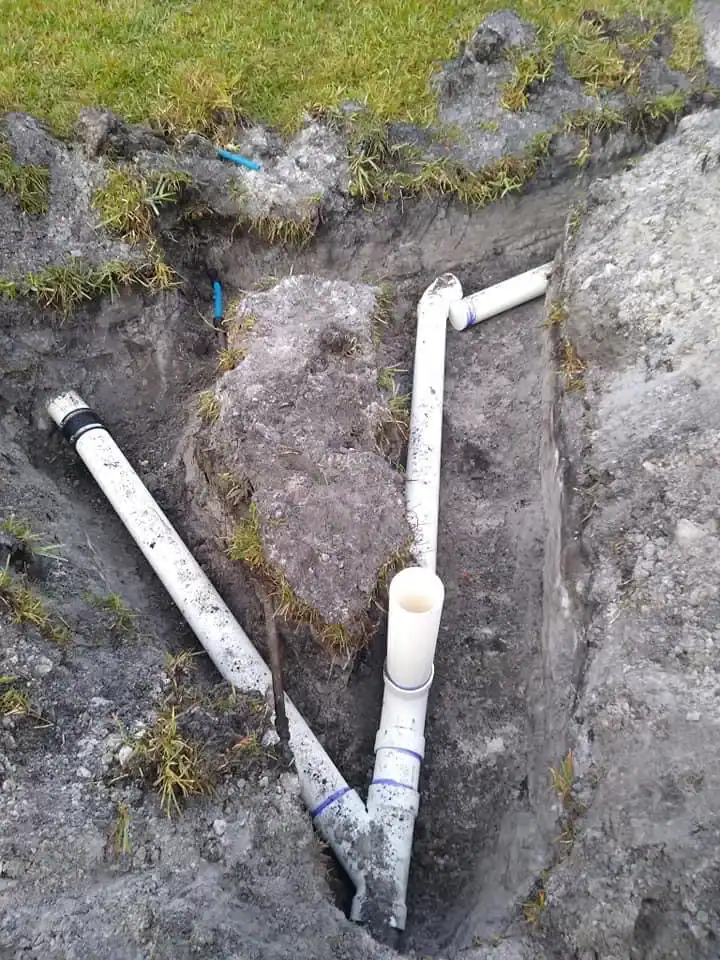 Two white PVC pipes in a trench were installed around a large buried rock by a skilled plumber Brevard County, FL. The pipes are bent to avoid the obstacle, with dug-up ground and some grass visible along the edges.