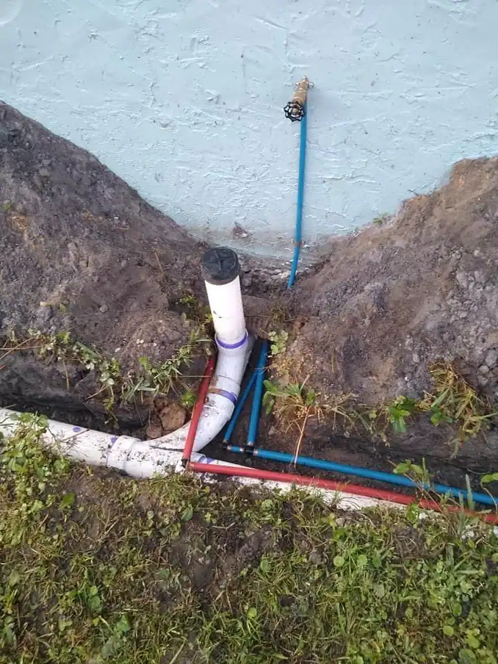 A patch of ground with dug-out soil reveals exposed blue, red, and white pipes arranged around a PVC pipe emerging vertically near a blue wall with a faucet above—typical work for a plumber in Brevard County, FL. Grass surrounds the dug-up area.