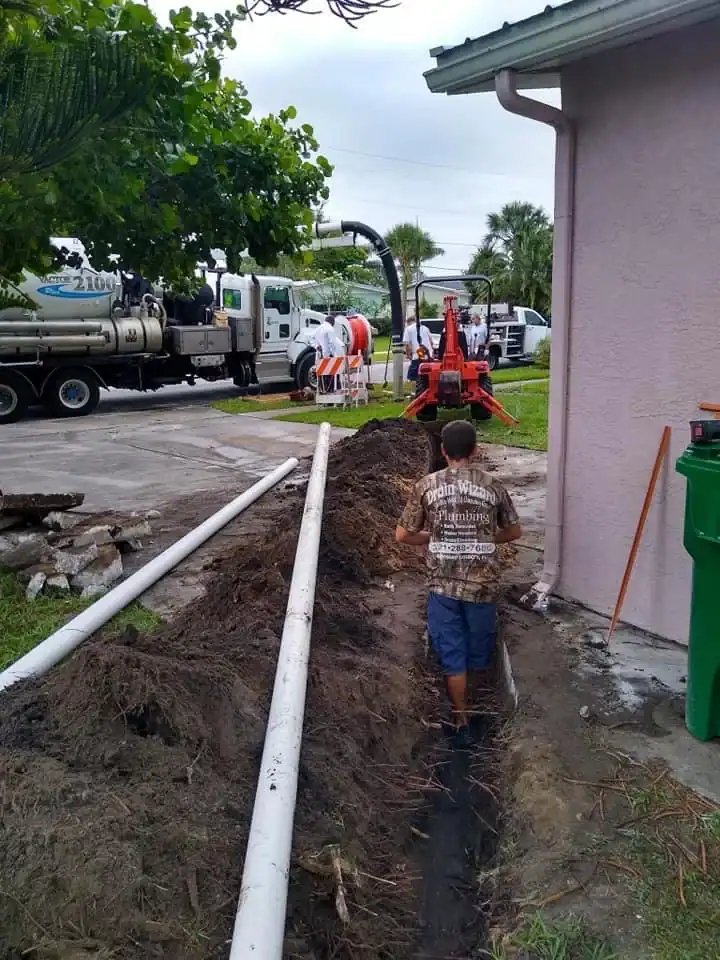 A young boy stands in a trench beside a house in FL while workers and utility trucks, led by a plumber Brevard County team, conduct sewer or pipe work nearby. White pipes lie parallel to the trench, with equipment in the background.