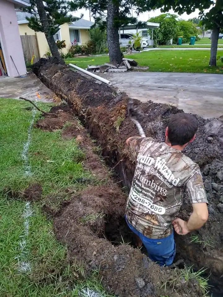 A plumber in Brevard County, FL, wearing a muddy shirt stands in a deep trench, repairing or installing a pipe in the yard of a residential neighborhood with grass, trees, and houses visible in the background.
