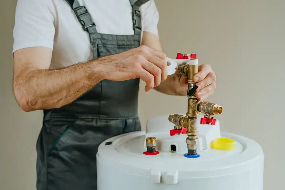 A plumber in Brevard County, FL, wearing grey overalls adjusts plumbing valves on top of a white hot water heater, performing maintenance or installation work.