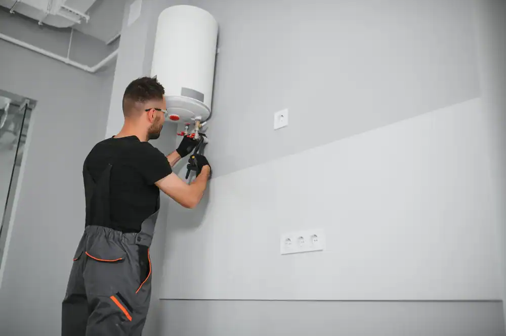 A man in work clothes and gloves is installing or repairing a white water heater mounted on a gray wall inside a modern room.