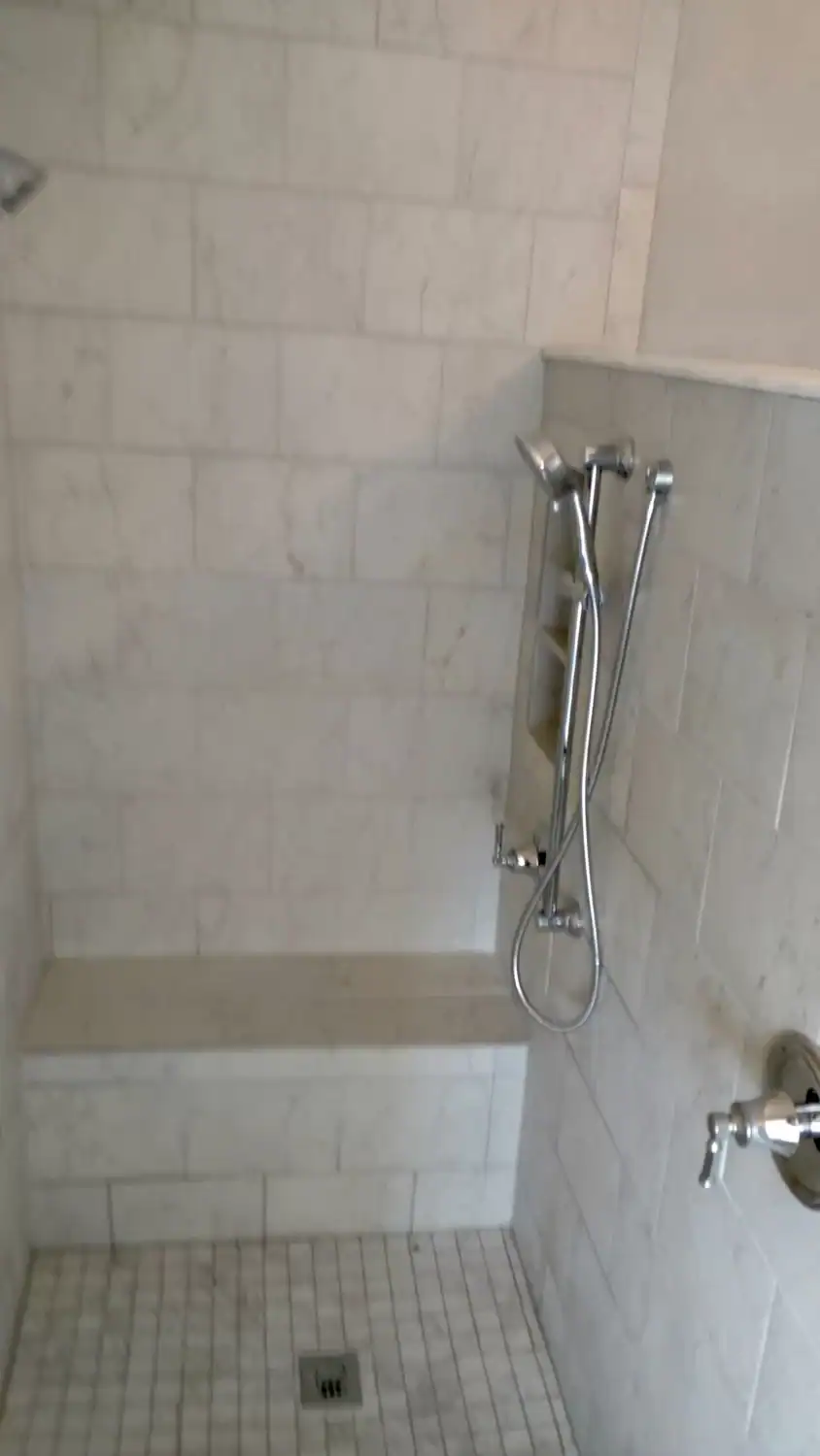 A tiled shower with a built-in bench, a handheld showerhead mounted on the wall, and a drain on the floor. The tiles are light-colored and rectangular.