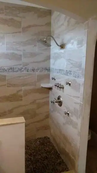 A walk-in shower with beige tiled walls, a built-in shelf, decorative tile trim, and a dark pebble stone floor. Multiple silver shower controls and a showerhead are expertly installed by a trusted plumber Brevard County, FL.