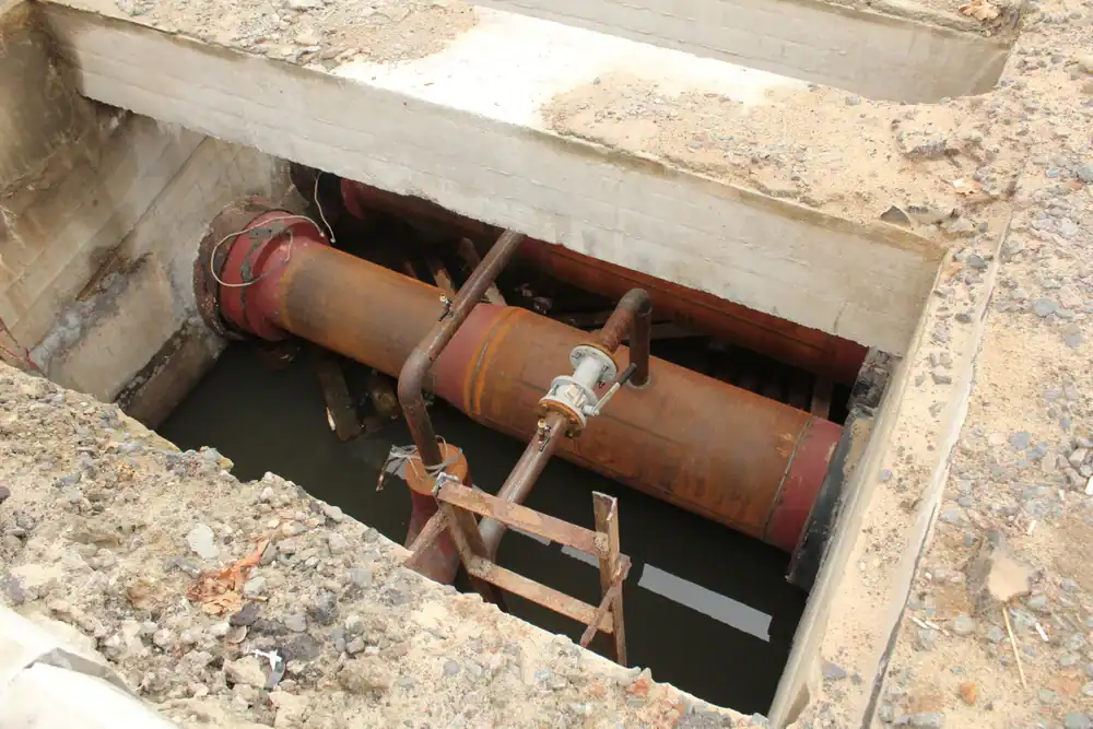 A large rusty pipe runs through a concrete trench partially filled with water, surrounded by soil and construction debris, with a metal ladder leaning into the trench—typical of what a plumber in Brevard County, FL might encounter on the job.
