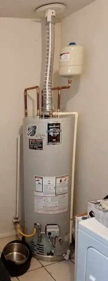 A water heater with attached pipes and a vent is installed in a corner of a room by a plumber in Brevard County, FL. A small expansion tank is mounted above it, with a washing machine and pet water bowl nearby.