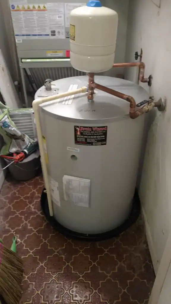 A water heater with pipes connected at the top sits in a small utility room with patterned floor tiles. Nearby are cleaning supplies, a bucket, and a broom leaning against the wall.