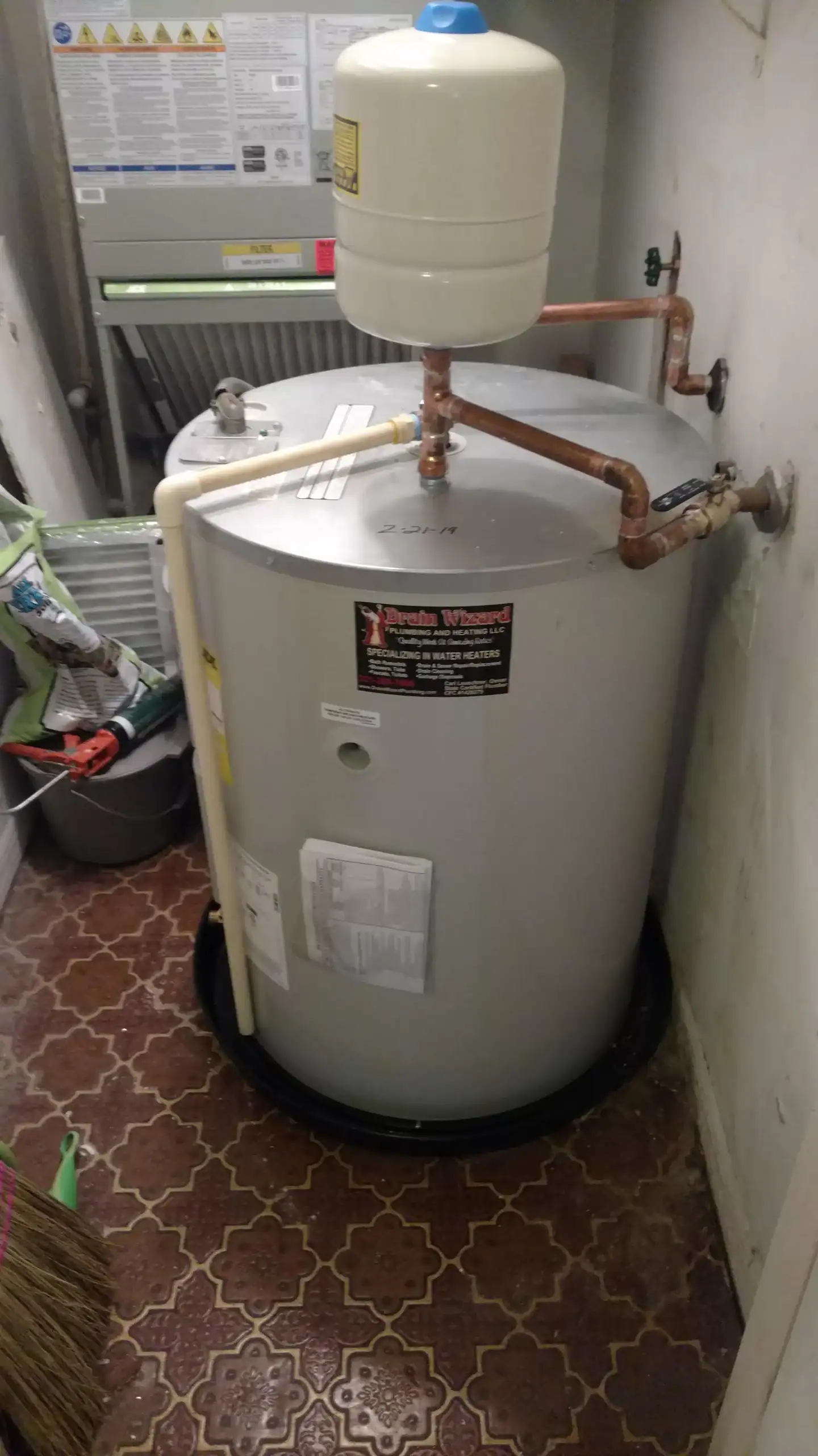 A large, cylindrical water heater with pipes and a pressure tank is installed in a FL utility room with cleaning supplies and patterned tile floor. Warning labels and paperwork are attached, suggesting recent work by a plumber Brevard County residents trust.