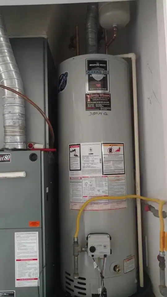 A tall, cylindrical water heater with warning labels and pipes connected, installed next to an HVAC unit inside a utility closet—FL plumber Brevard County details include yellow, silver, and copper pipes along with ventilation ducts above.