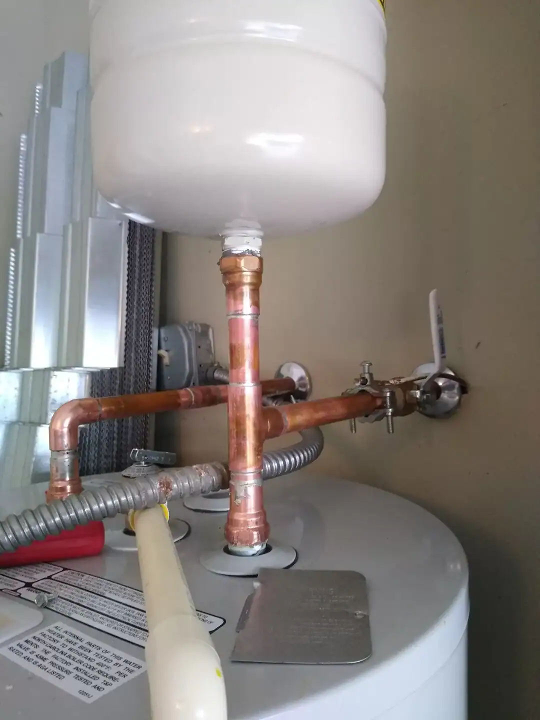 A close-up view of copper piping connected to a white water heater—with an attached expansion tank mounted above—set against a beige wall, as expertly installed by a plumber Brevard County, FL.