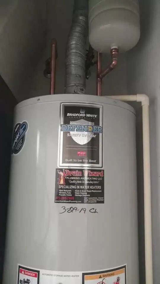 A Bradford White Defender System water heater with attached pipes and an expansion tank is installed in a utility space by a plumber Brevard County, FL. Several stickers and a handwritten “389-19 CL” are visible on the water heater.