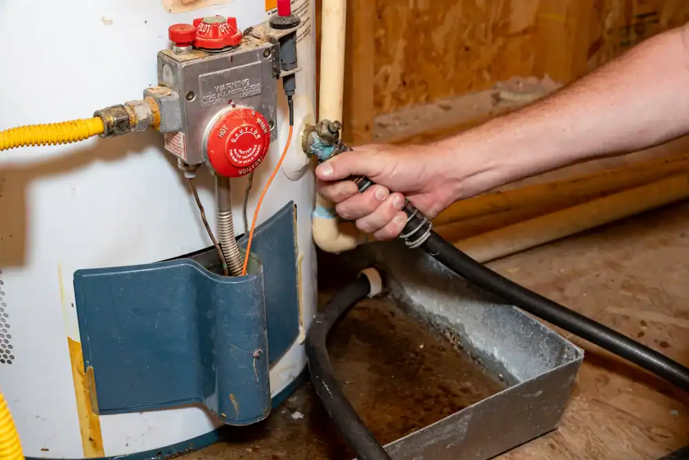 A plumber in Brevard County, FL, holds a hose connected to the drain valve at the bottom of a water heater, with a pan underneath to catch water. The water heater is located in a utility room.
