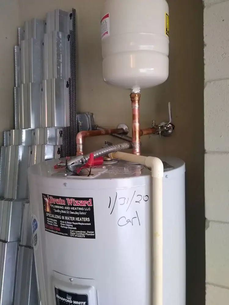 A water heater with copper pipes and a white expansion tank mounted above it, installed in a garage by a plumber Brevard County, FL. The date "1/31/20" and the name "Carl" are handwritten on the heater's surface.