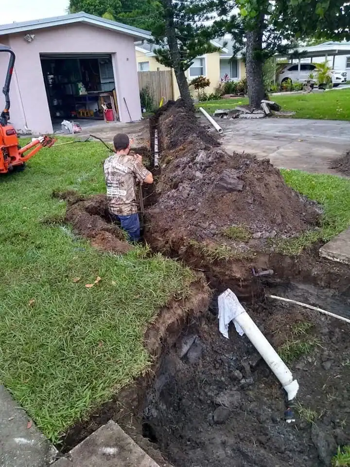 A plumber in Brevard County, FL digs a deep trench in a grassy yard near a house, exposing a white underground pipe. Dirt piles and a small excavator are nearby, with a garage and trees in the background.