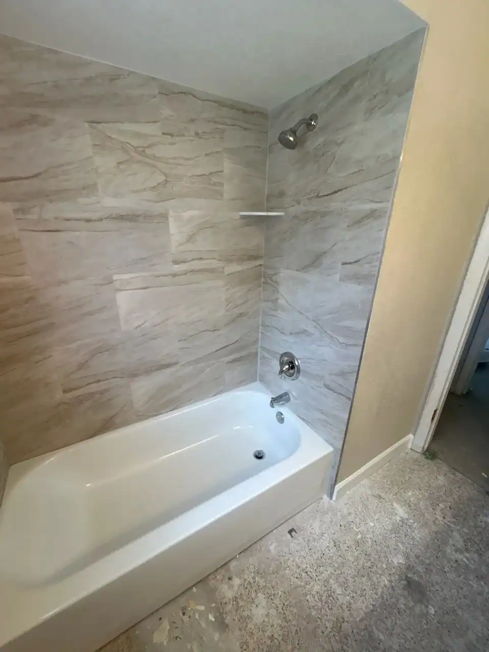 A bathroom with a white bathtub and gray marble-patterned wall tiles. The fixtures include a showerhead, faucet, and a small corner shelf. The floor appears unfinished, with exposed subfloor and scattered debris.